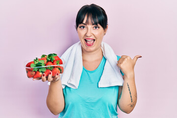 Young hispanic woman wearing sportswear holding strawberries pointing thumb up to the side smiling happy with open mouth