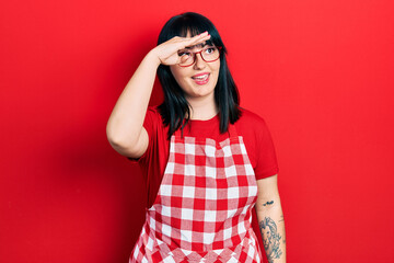 Young hispanic woman wearing cook apron and glasses very happy and smiling looking far away with hand over head. searching concept.