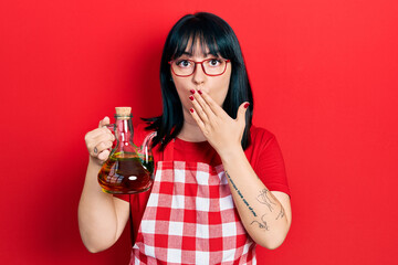 Young hispanic woman wearing cook apron holding olive oil can covering mouth with hand, shocked and afraid for mistake. surprised expression