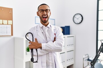 African american doctor man holding stethoscope at the clinic angry and mad screaming frustrated and furious, shouting with anger looking up.