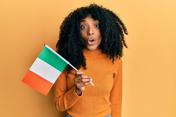 African american woman with afro hair holding ireland flag scared and amazed with open mouth for...