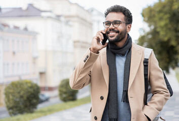 Young handsome student man using smartphone. Smiling joyful guy autumn portrait. Cheerful businessman wearing warm clothes talking by mobile phone in a city