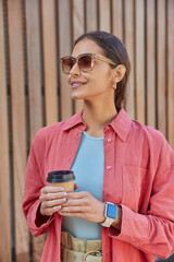 Pensive female model has coffee break after work poses with disposable cup of aromatic beverage tasty cappuccino wears sunglasses stylish outfit waits for someone outdoor. Lifestyle concept.
