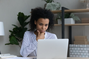 Analyzing information. Pensive mixed race female office worker read analytic report from laptop screen consider on project solution. Black business lady sit by pc think on partner offer make decision