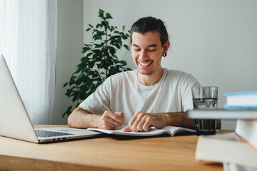 Cheerful student making notes from laptop while online learning. Young man studying at home and prepare for pass exam to college