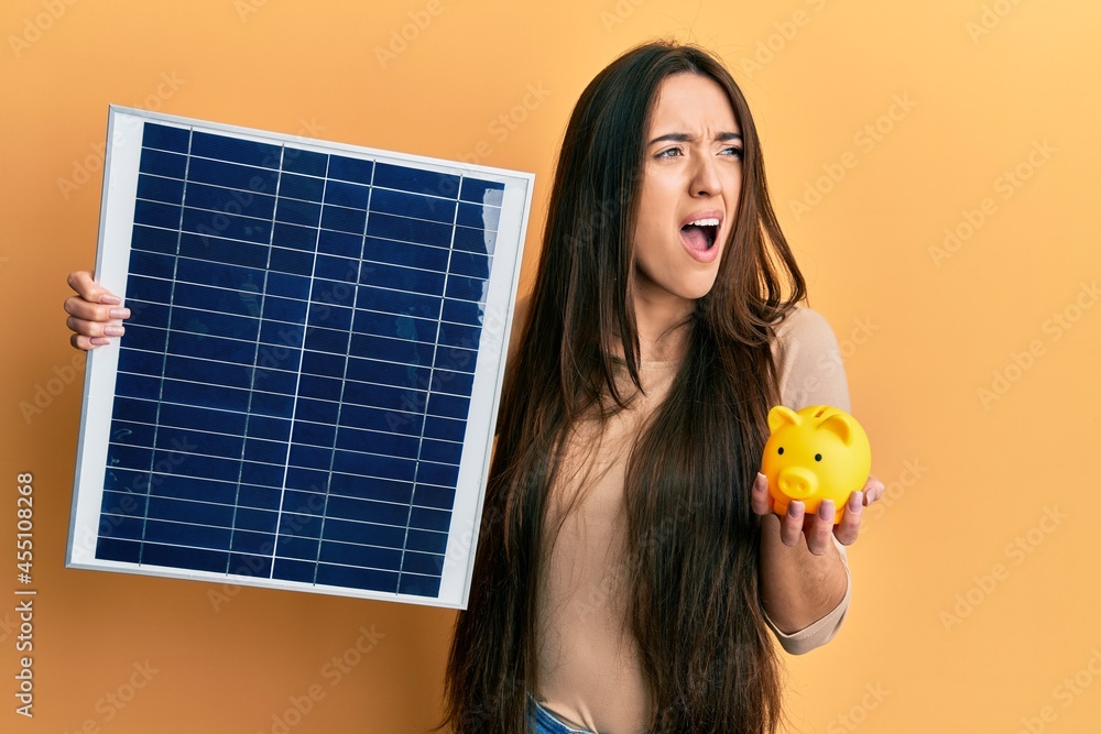 Poster young hispanic girl holding photovoltaic solar panel and piggy bank angry and mad screaming frustrat