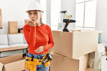 Middle age grey-haired woman wearing hardhat standing at new home looking at the camera blowing a kiss with hand on air being lovely and sexy. love expression.