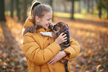 Naklejka na ściany i meble Pretty blonde girl in yellow jacket holding on hand dog and smiling on park background. Concept of child playing with dog in park.