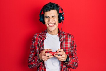 Young hispanic man playing video game holding controller winking looking at the camera with sexy...