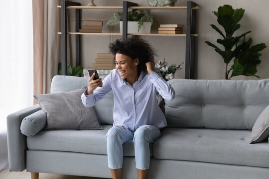 Excited News. Overjoyed Cheerful Mixed Race Female Read Amazing Message On Cell Of Getting Money Prize. Lucky Black Woman Celebrate Success In Lottery Betting Look At Phone Screen Sit On Couch At Home