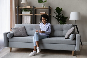 Full length shot of casual black female student wear earphones sit on couch at modern living room talk by videocall using pad computer. Young biracial lady speak in video chat in app for tablet device