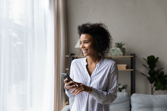 Happy mixed race gen z female stand at living room hold mobile phone look outdoors wait for beloved person call think about future dating. Cheerful young black woman using cell near window. Copy space - Powered by Adobe