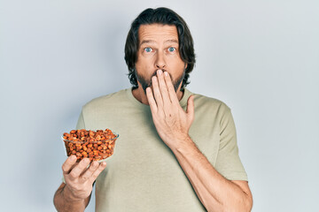 Middle age caucasian man holding peanuts covering mouth with hand, shocked and afraid for mistake....