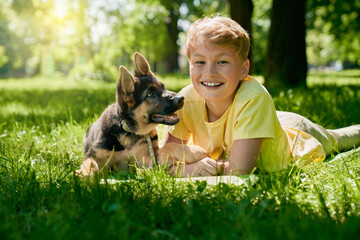 Cheerful teenage boy and german shepherd puppy lying on green grass during sunny days. Happy child...