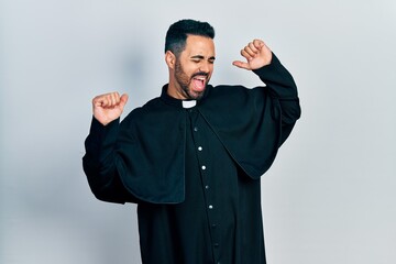 Handsome hispanic man with beard wearing catholic priest robe dancing happy and cheerful, smiling moving casual and confident listening to music