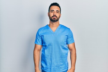 Handsome hispanic man with beard wearing blue male nurse uniform relaxed with serious expression on...
