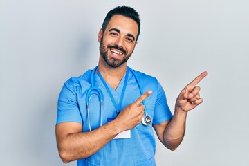 Handsome hispanic man with beard wearing doctor uniform smiling and looking at the camera pointing...