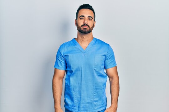 Handsome Hispanic Man With Beard Wearing Blue Male Nurse Uniform Relaxed With Serious Expression On Face. Simple And Natural Looking At The Camera.