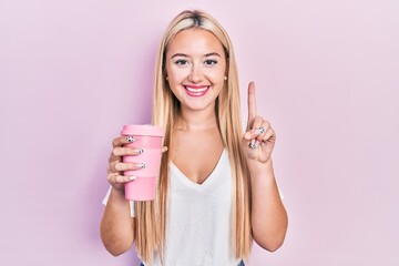 Young blonde girl holding coffee smiling with an idea or question pointing finger with happy face, number one
