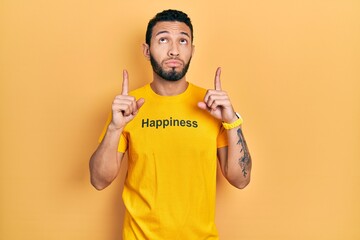 Hispanic man with beard wearing t shirt with happiness word message pointing up looking sad and...