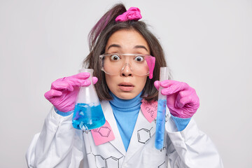 Stunned female scientist examines blue solution in glassware cultivates vaccine against virus wears protective glasses medical coat poses indoor. Professional medical development researcher at lab