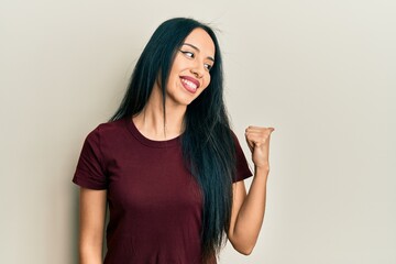 Young hispanic girl wearing casual t shirt pointing thumb up to the side smiling happy with open mouth