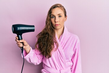 Young blonde woman wearing bathrobe using hair dryer thinking attitude and sober expression looking self confident