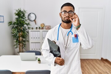 Young indian man wearing doctor uniform and stethoscope mouth and lips shut as zip with fingers....