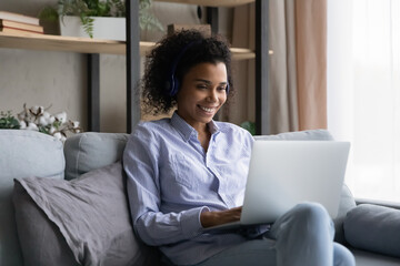 Easy learning. Smiling young mixed race lady in headphones sit on couch search information using laptop watch webinar video lesson. Positive black woman use online education service to improve skills