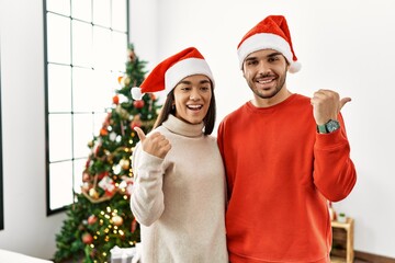 Fototapeta premium Young hispanic couple standing by christmas tree smiling with happy face looking and pointing to the side with thumb up.