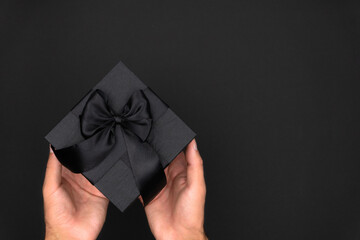 Black gift box with black satin ribbon in male hands on a black background.