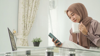 beautiful woman in hijab working from home looking for good ideas on her smartphone while enjoying...