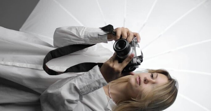 Portrait of a beautiful young woman photographer in a white shirt with long blond hair, making shots, looking into the camera's video lens and smiling. Vertical video. Professions, hobbies, the arts.