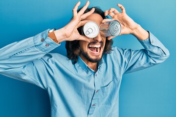 Young hispanic man holding canned food over eyes angry and mad screaming frustrated and furious, shouting with anger. rage and aggressive concept. - obrazy, fototapety, plakaty