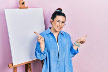 Young hispanic woman standing by painter easel stand smiling confident pointing with fingers to different directions. copy space for advertisement