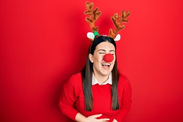 Young hispanic woman wearing deer christmas hat and red nose smiling and laughing hard out loud...
