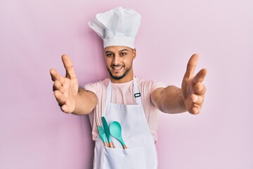 Young arab man wearing professional cook apron and hat looking at the camera smiling with open arms for hug. cheerful expression embracing happiness.