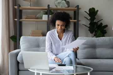 Paying insurance fee. Smiling mixed race woman client do paperwork at home office calculate regular payment by loan mortgage contract. Young biracial lady manage financial documents pay using laptop