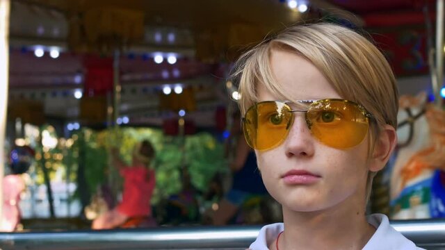 A frustrated teenage guy stands by the carousels, he cannot ride the carousels because he is punished for bad behavior. The boy is blond in yellow glasses.