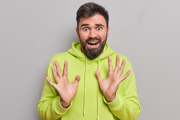 Fototapeta premium Scared panicking bearded man looks frightened has phobias raises palm tries to protect himself from something awful wears casual hoodie looks desperate and devastated isolated over grey background