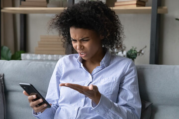 Phone problem. Angry black teen lady sit on sofa has unpleasant video conversation on cellphone...