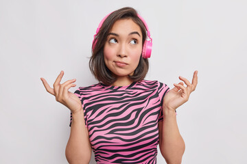 Maybe yes or no. Hesitant young Asian woman shrugs shoulders has clueless expression makes choice daydreams while listening music wears casual striped t shirt isolated over white background.