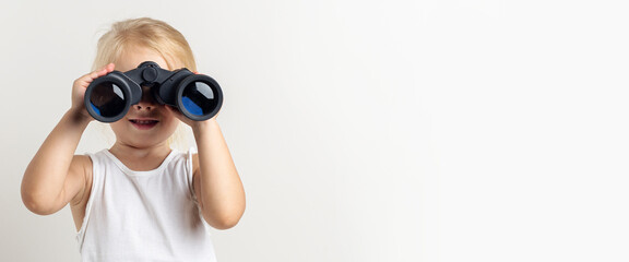 Smiling blonde child looks through binoculars on a light background in the studio. Banner