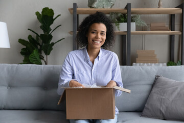 Moving day. Portrait of excited young biracial woman realty buyer renter sit on sofa hold big box...