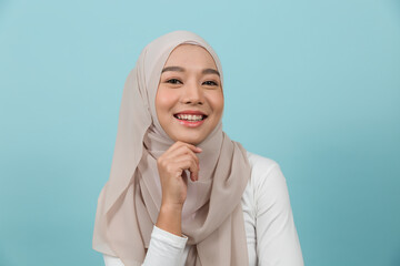 Portrait of smiling Asian young Muslim woman in hijab head scarf standing against blue colour background