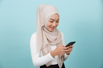 Portrait of smiling Asian young Muslim woman in hijab head scarf standing and using smart phone against blue colour background
