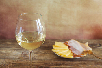 A glass of white wine stands on a wooden table, behind a plate of cheese and bacon.