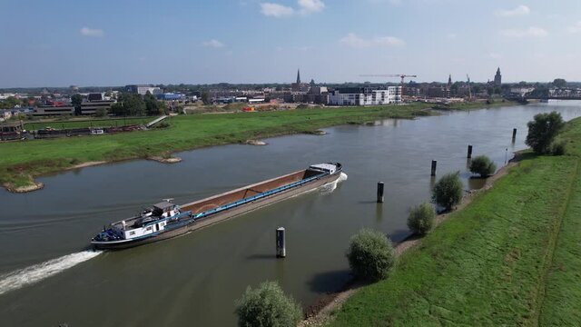 Aerial descend showing empty container vessel passing along river IJssel with construction site and Hanseatic tower town Zutphen in the background and floodplains in the foreground on a sunny day