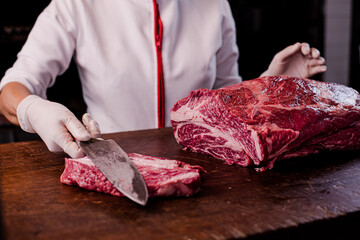 Chef cuts wagyu chuck steak on a large wooden cutting board. There is a grill in the background.