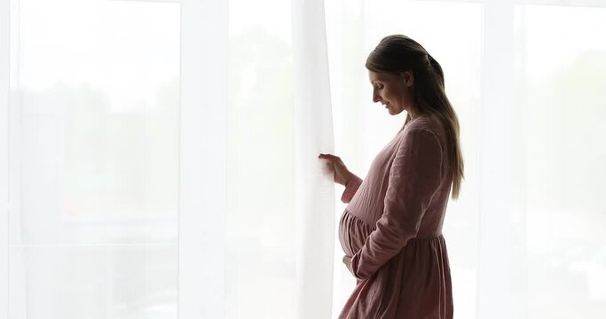 Side view young attractive calm 35s woman in long pink dress standing alone indoors touch her big pregnant belly looking out window into distance, in anticipation of childbearing. Motherhood concept